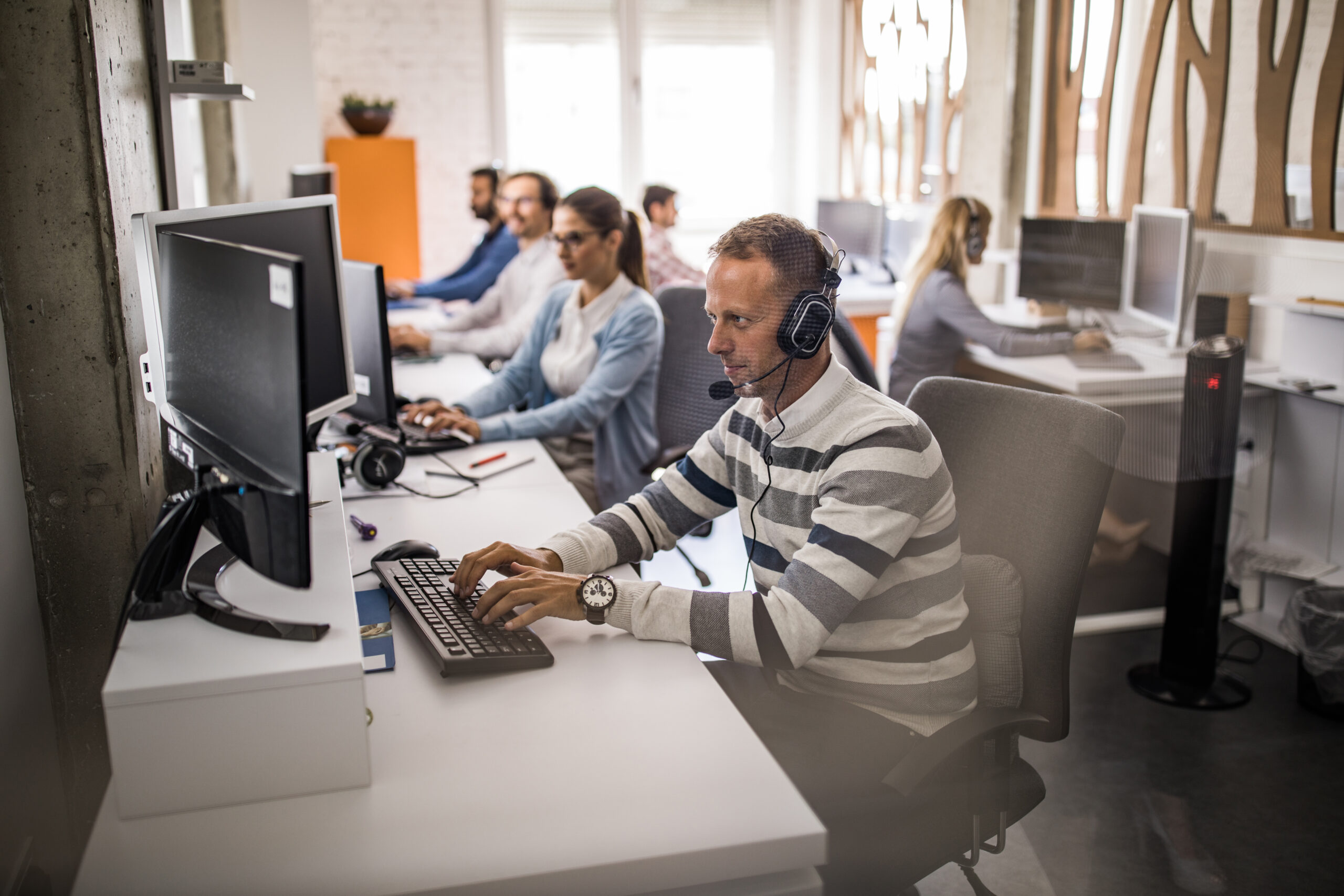 Customer service representative working on desktop PC in the office. Young man working on desktop PC at call center with his colleagues. The view is through glass.